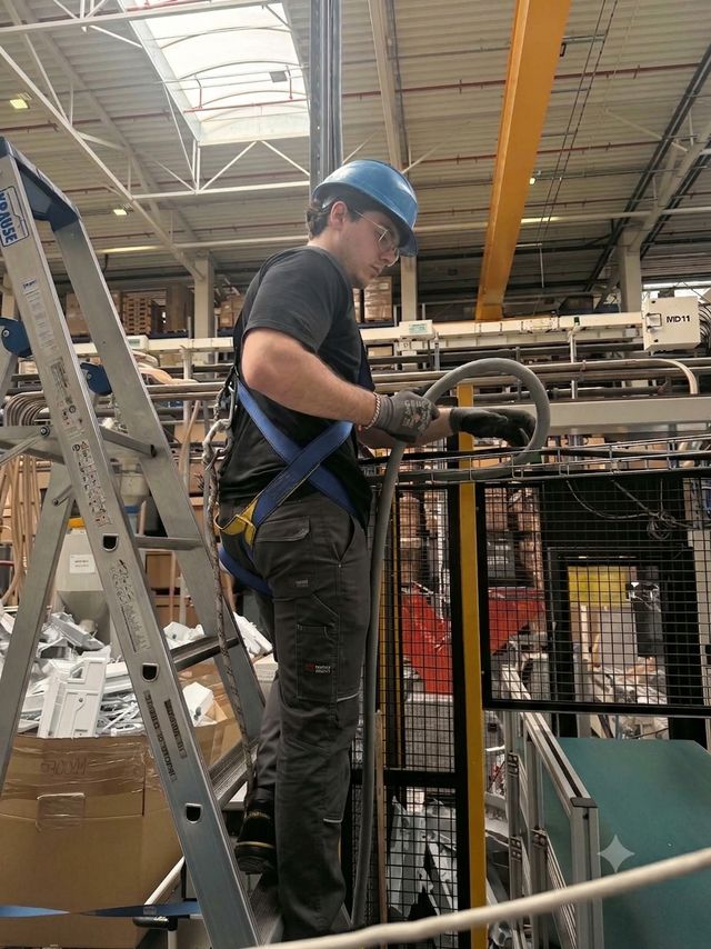 Technik w kasku i szelkach bezpieczeństwa prowadzi przewód zasilający na wysokości w hali produkcyjnej
