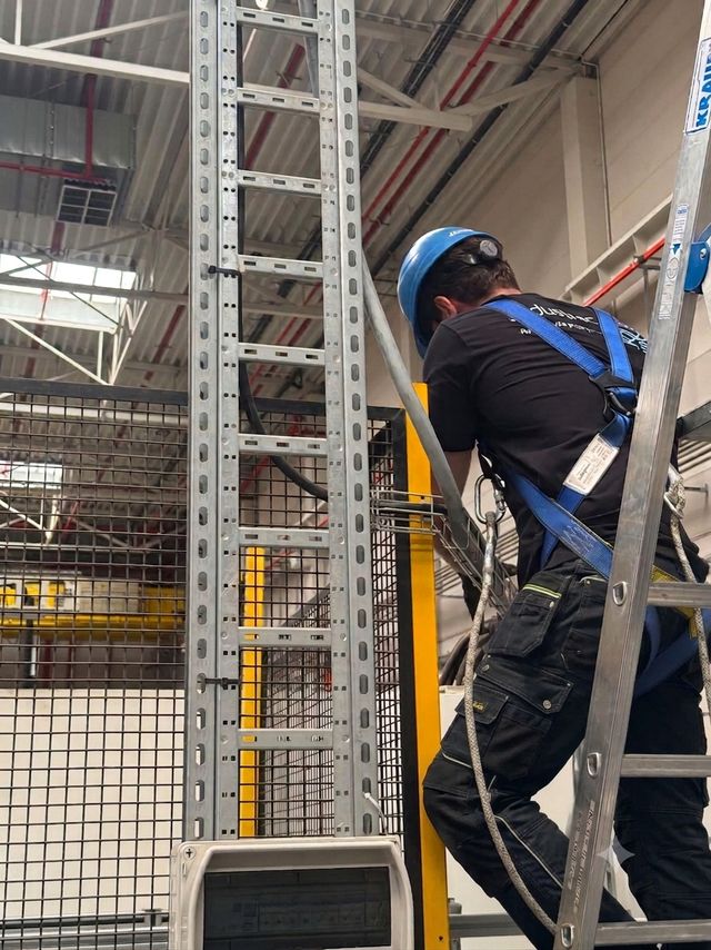 Technik w kasku i szelkach bezpieczeństwa montuje trasę kablową przy wygrodzeniu maszyny